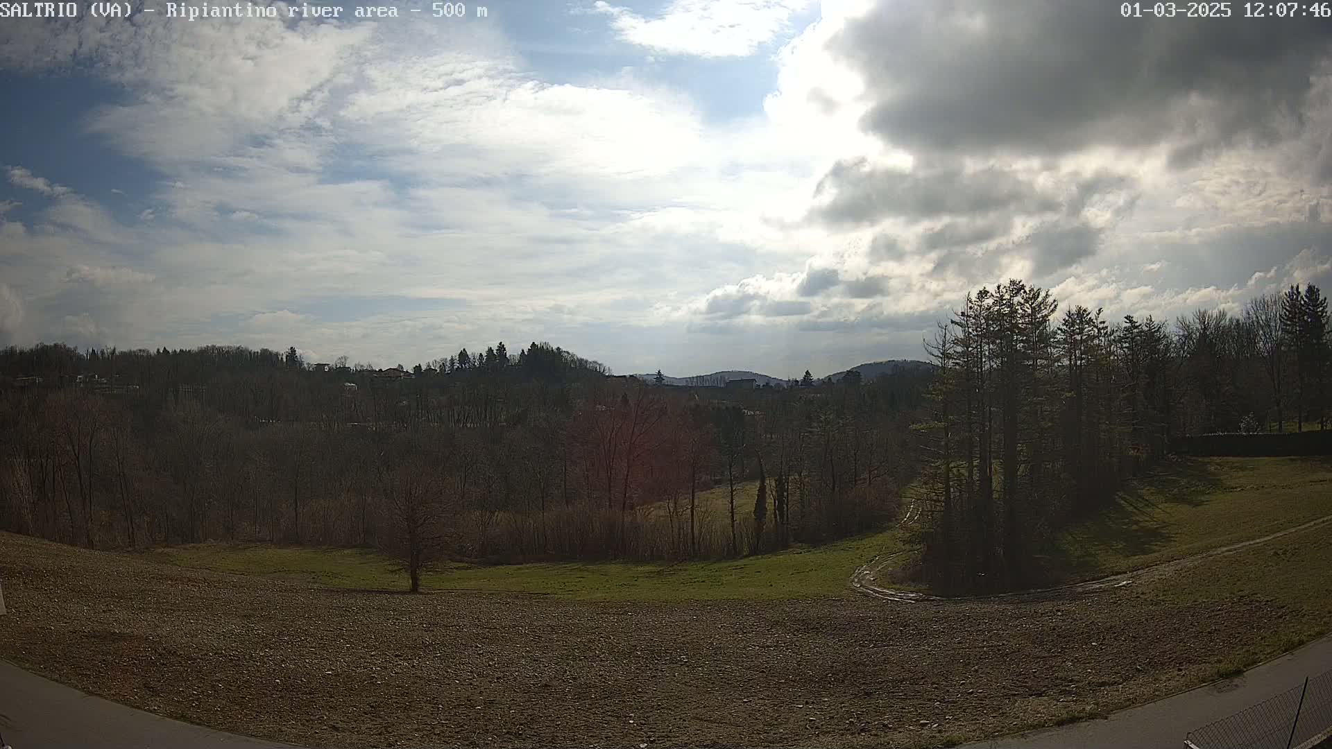 A partly cloudy sky overlooks a hilly landscape of bare trees and tilled fields.