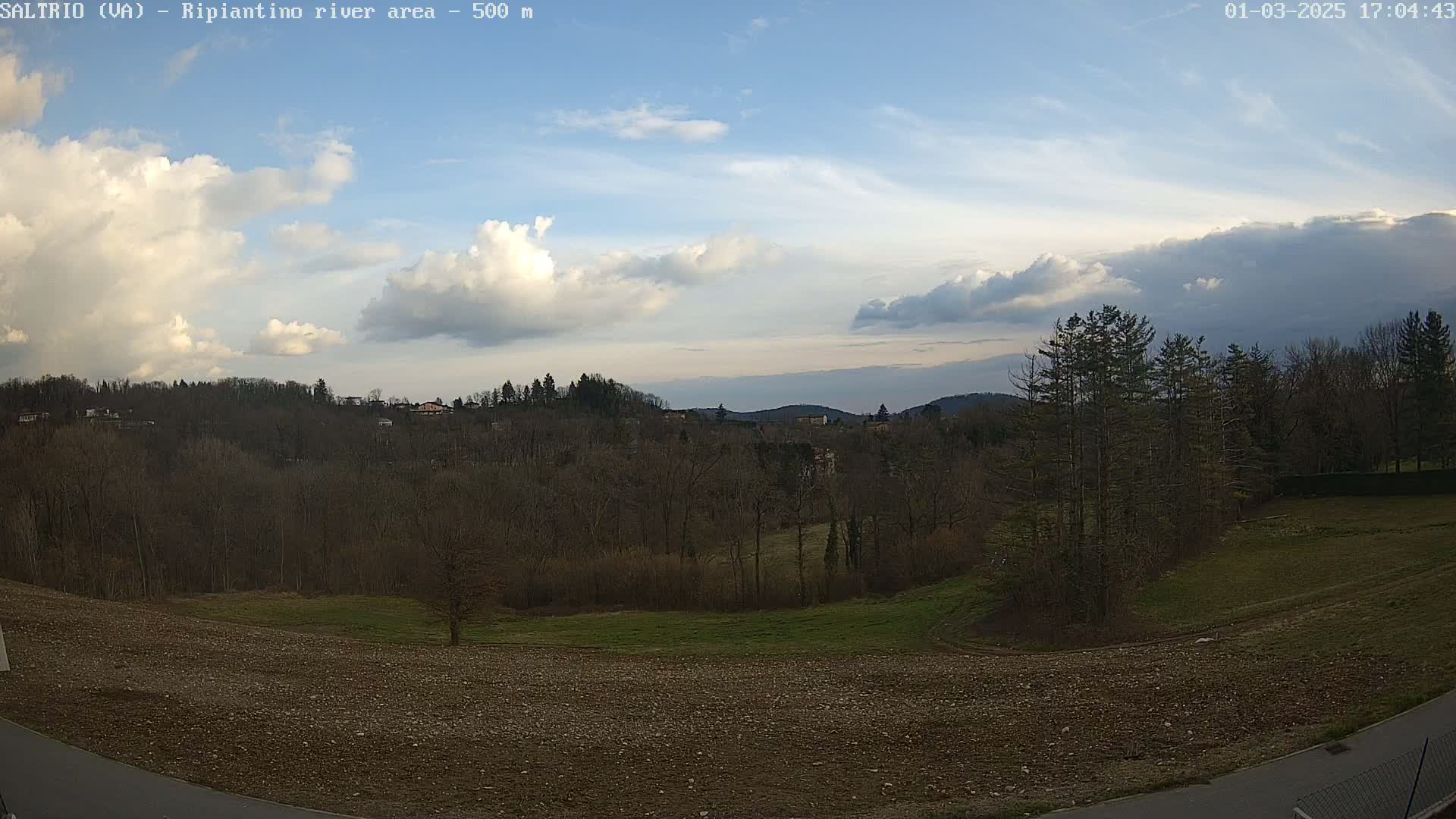 A mostly sunny landscape with partly cloudy skies shows bare winter trees and a plowed field in the foreground, overlooking a wooded hillside with distant mountains.