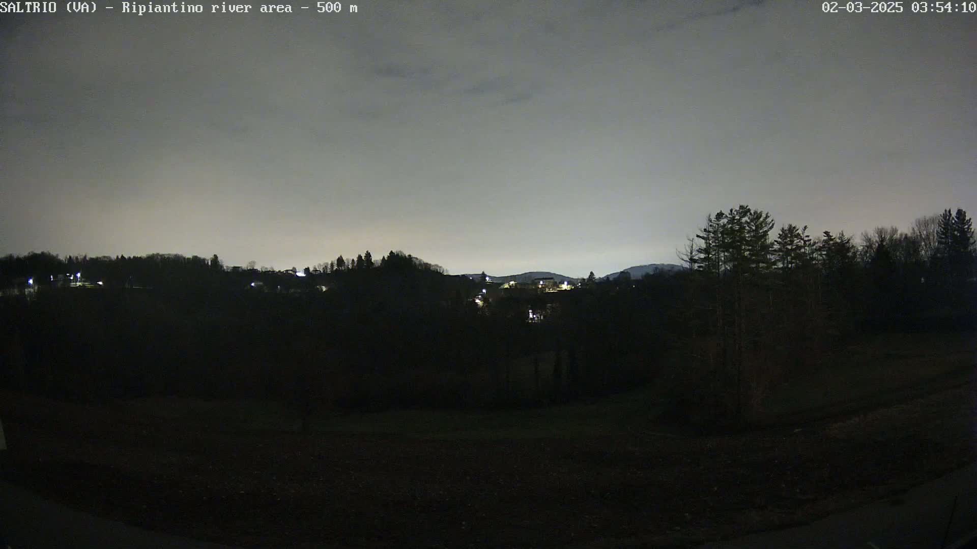 A nighttime view shows a dark, hilly landscape with sparsely lit buildings in the distance under a gray sky.