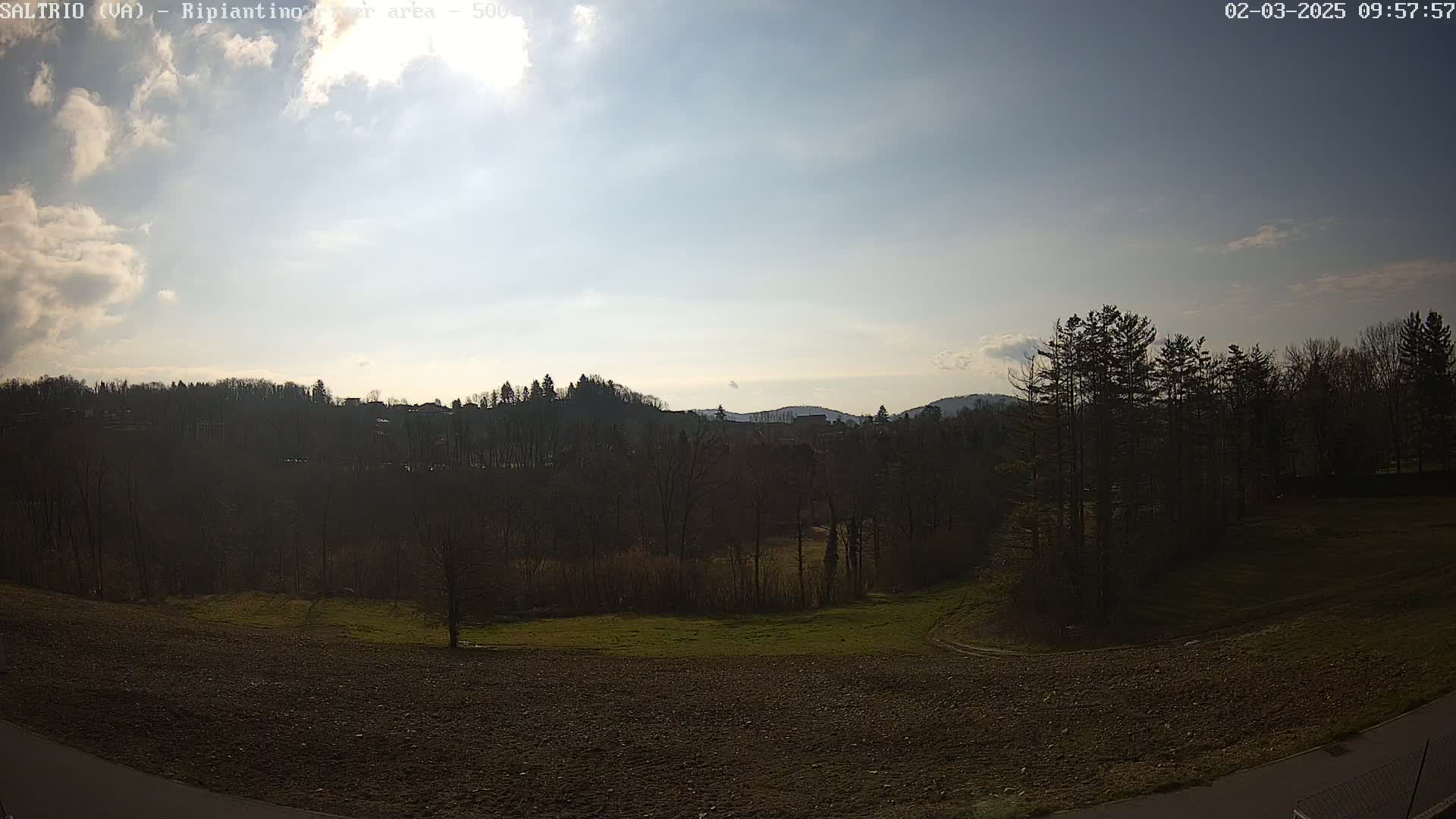 A mostly sunny day reveals a hilly landscape with a mix of bare trees and tilled fields, some buildings are visible in the distance.