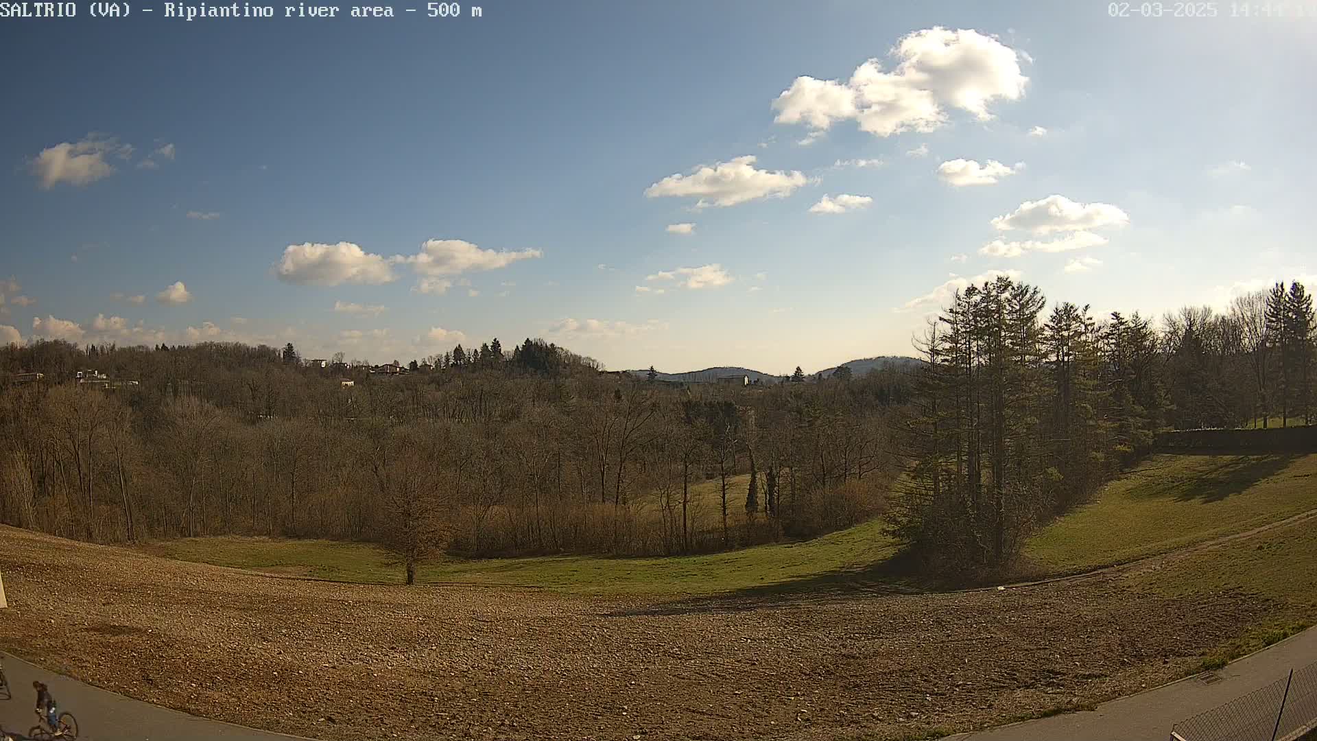 A partly sunny day reveals a hilly landscape with bare winter trees, a plowed field, and a cyclist on a paved path in the foreground.