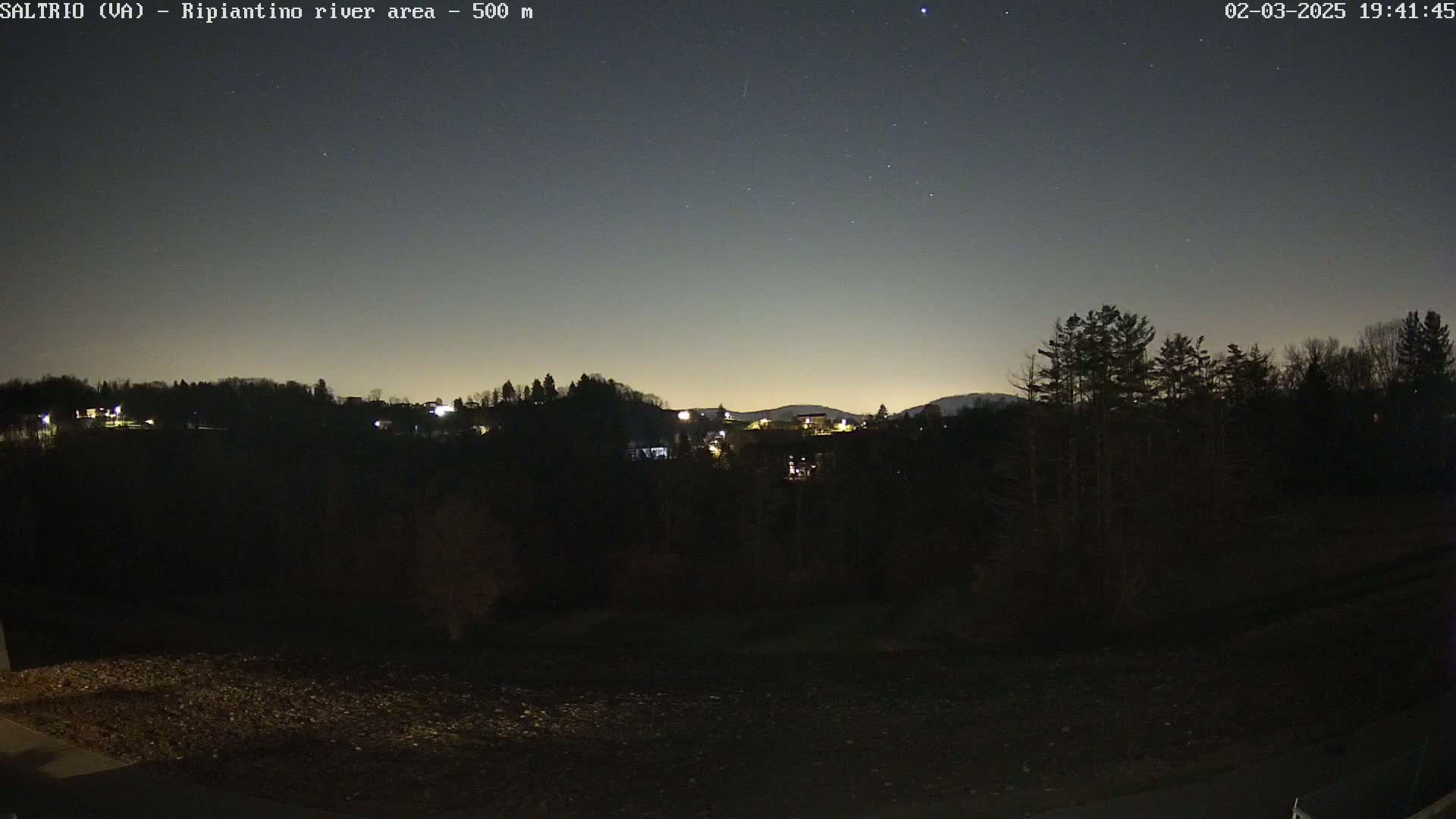 A nighttime view of a dark, tree-lined hill with a few distant lights, under a clear, starry sky.