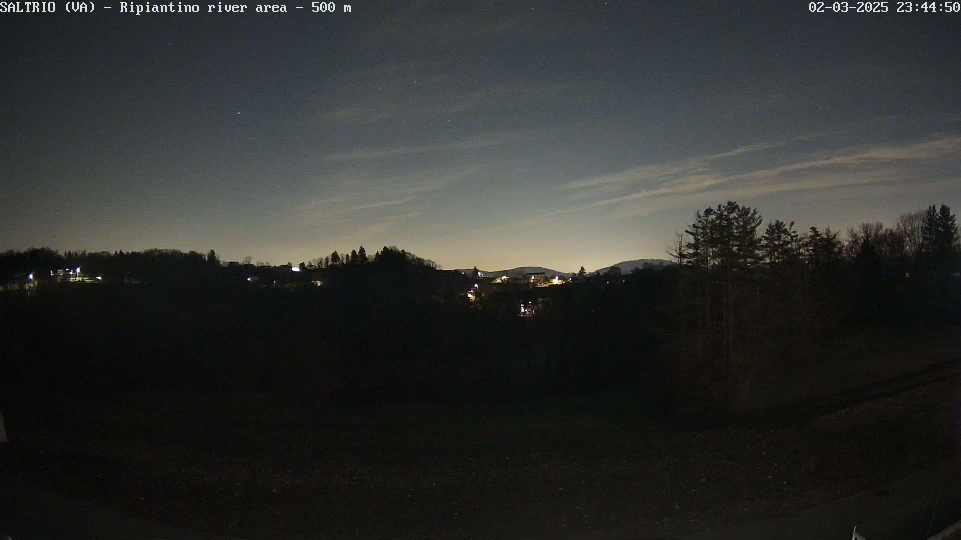 A nighttime view of a dark, silhouetted landscape with a few lights visible in the distance under a mostly clear sky with faint clouds.