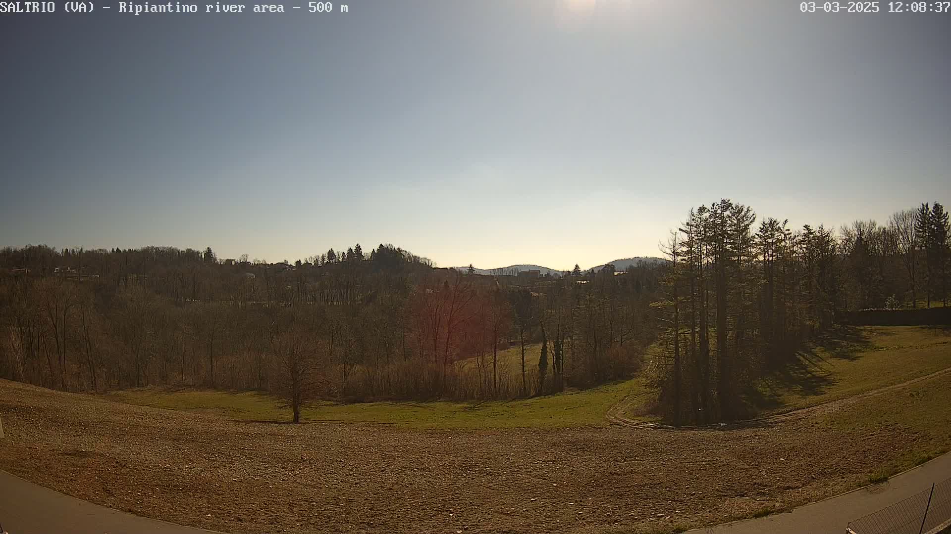A sunny day reveals a mostly barren field next to a line of leafless trees, beyond which lie more trees and a distant village nestled into hills.
