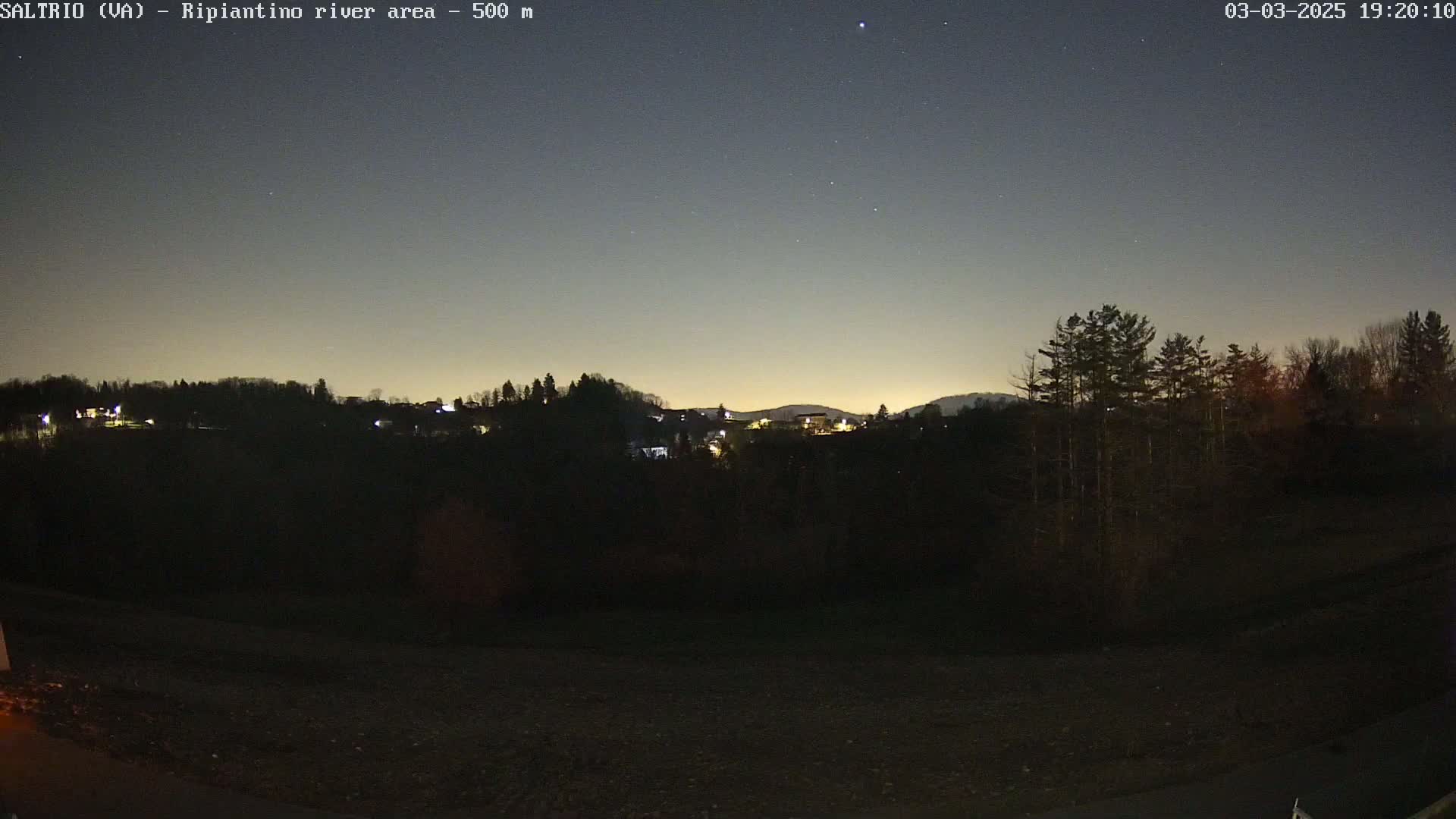 A nighttime view of a dimly lit town nestled in a valley, under a twilight sky with some stars visible.