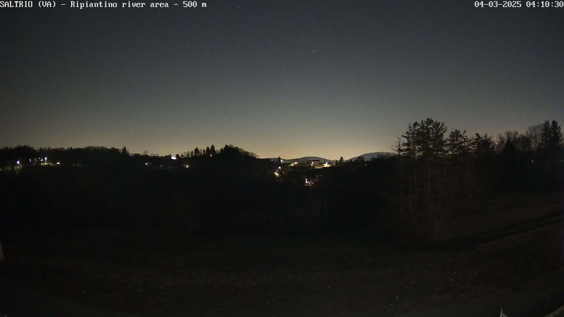 A nighttime view of a dark, hilly landscape with a few distant lights under a clear, starry sky.