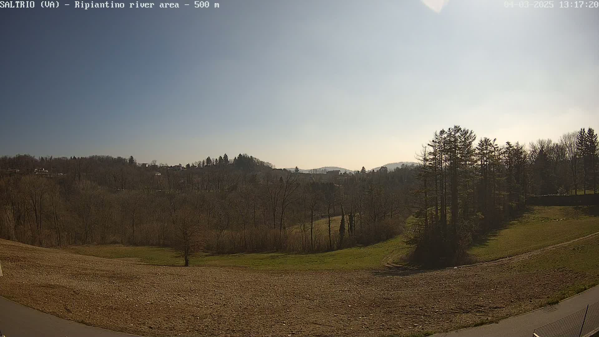 A mostly sunny day reveals a landscape of bare winter trees, a grassy field, and brown tilled earth, with distant hills in the background.