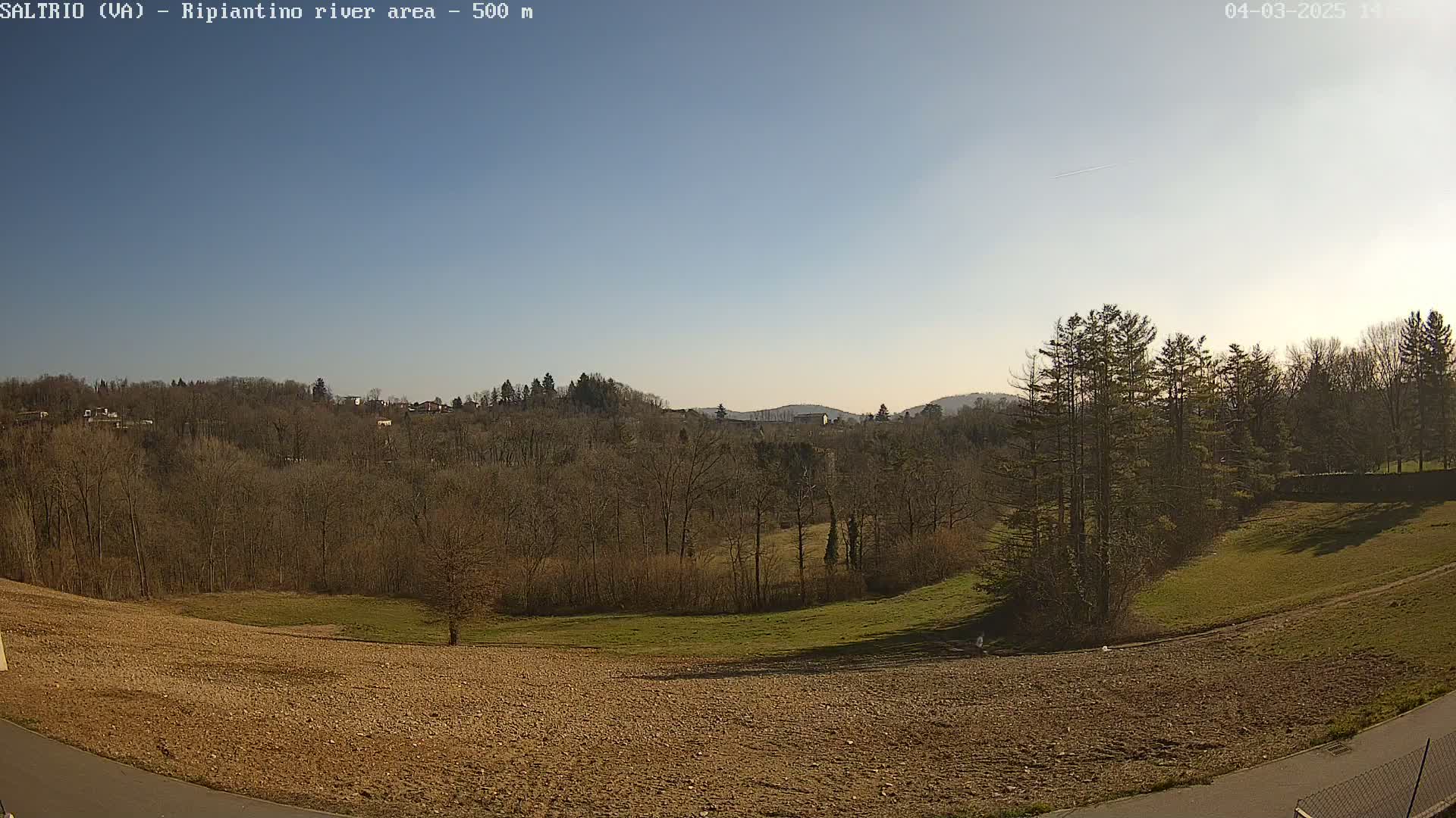 A mostly clear blue sky overlooks a brown and green landscape of barren trees, grassy fields, and tilled earth.