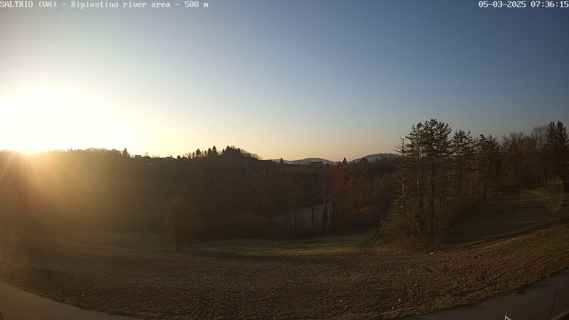 A sunrise illuminates a flat, plowed field, with a line of trees and hills in the background under a clear sky.