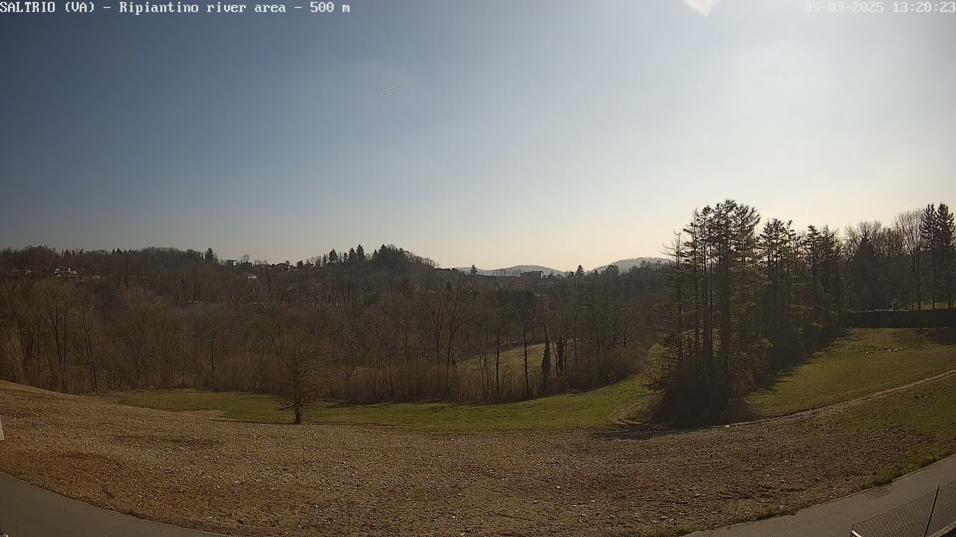 A mostly sunny day reveals a view of a plowed field, grassy areas, and a line of leafless trees leading to a hilly landscape in the distance.