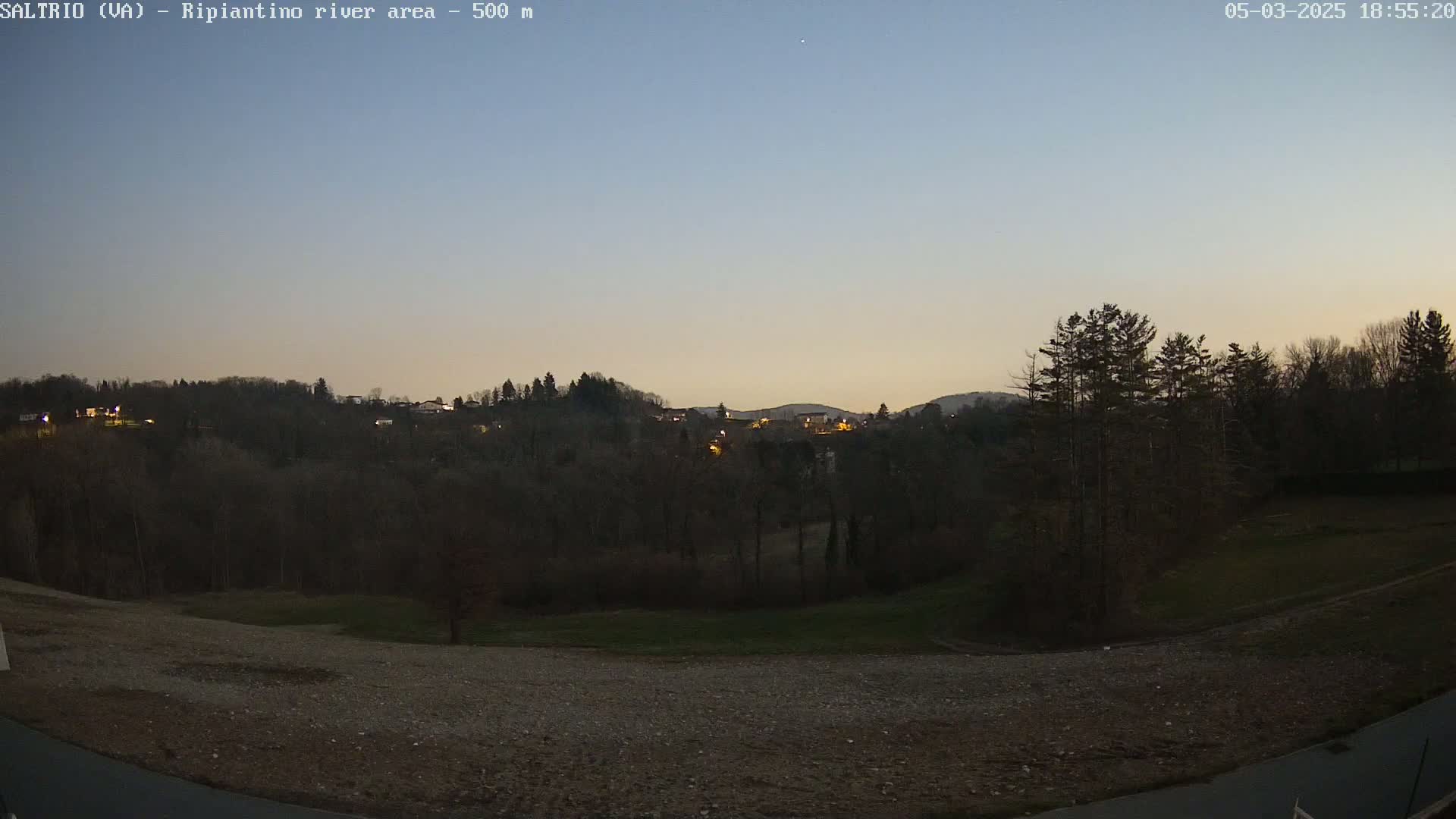 A twilight view of a dark, tree-covered hillside with a few lights visible, a flat, gravelly area in the foreground, and a clear, light-blue sky.