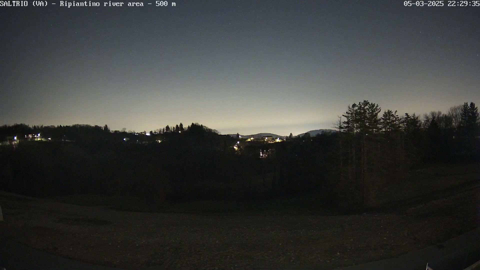 A nighttime view of a dark, tree-lined hillside with a few distant lights, under a mostly clear, twilight sky.