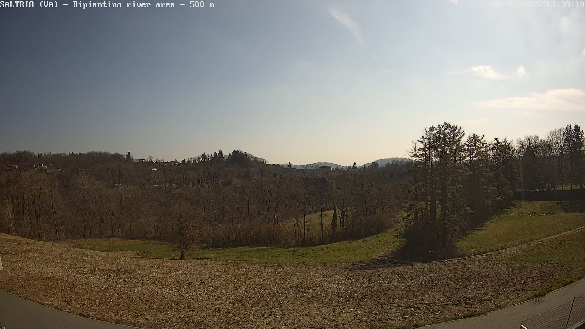 A sunny, mostly clear day reveals a view of a hilly landscape with bare winter trees, patches of green grass, and a brown, plowed field in the foreground.