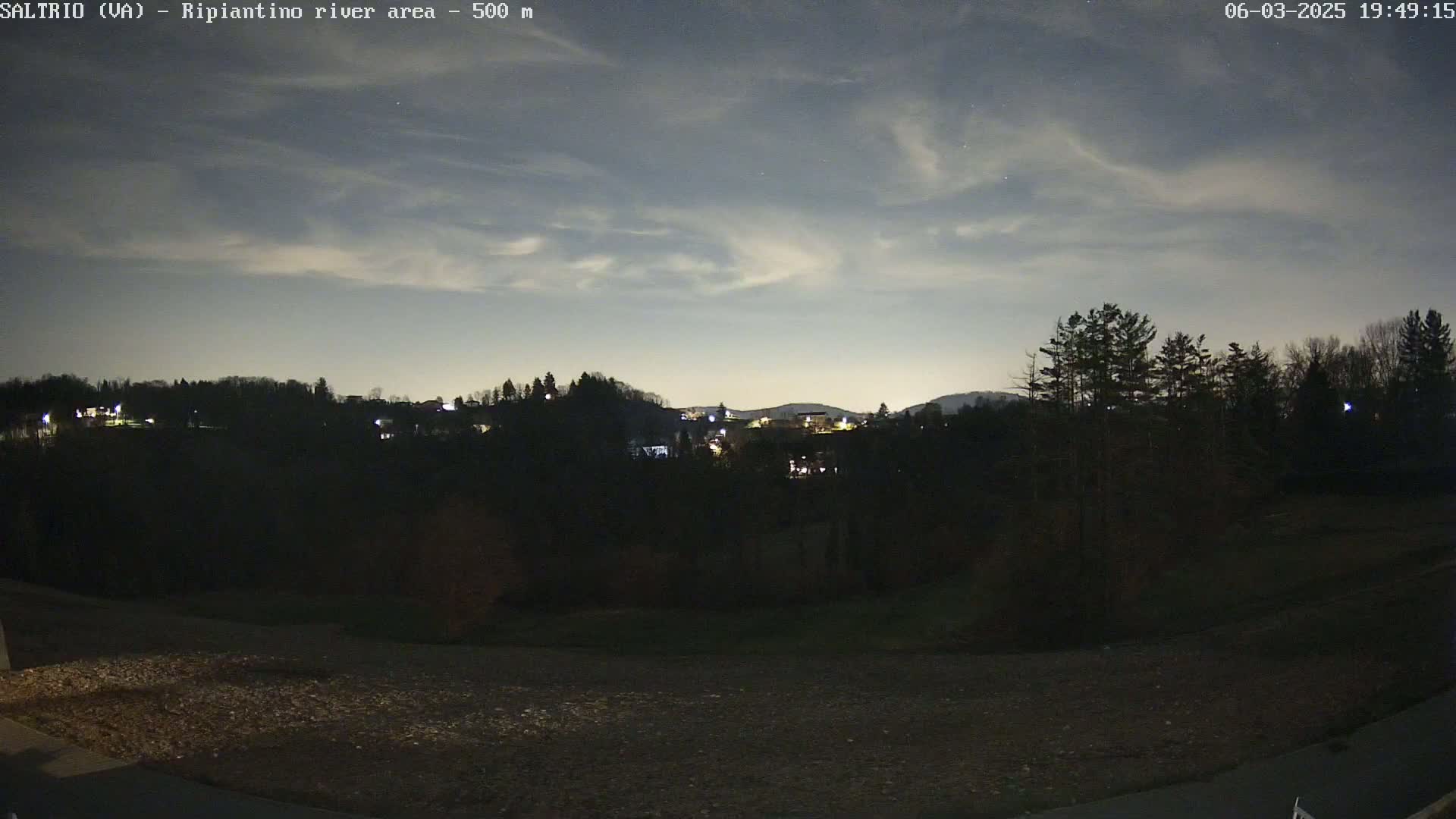 A nighttime view of a hilly landscape with a few scattered lights from buildings under a mostly cloudy sky.