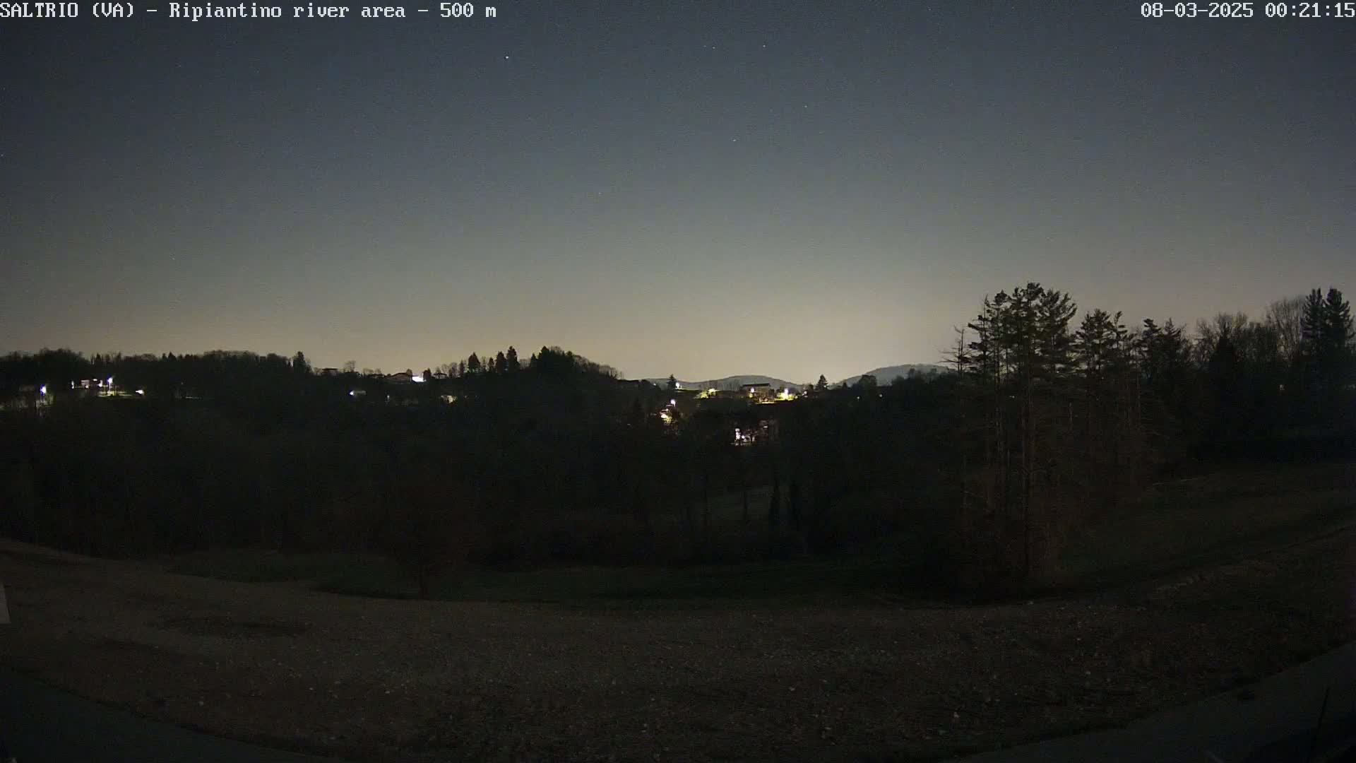 A nighttime view of a dark, treeline hillside with a few distant lights, under a clear, dark sky.