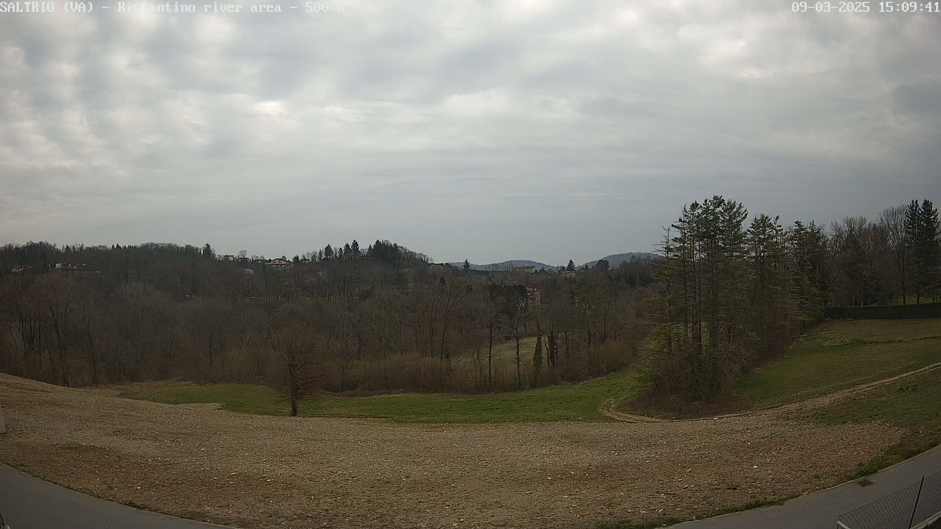 A mostly barren, brown field slopes down to a wooded area under a cloudy sky.