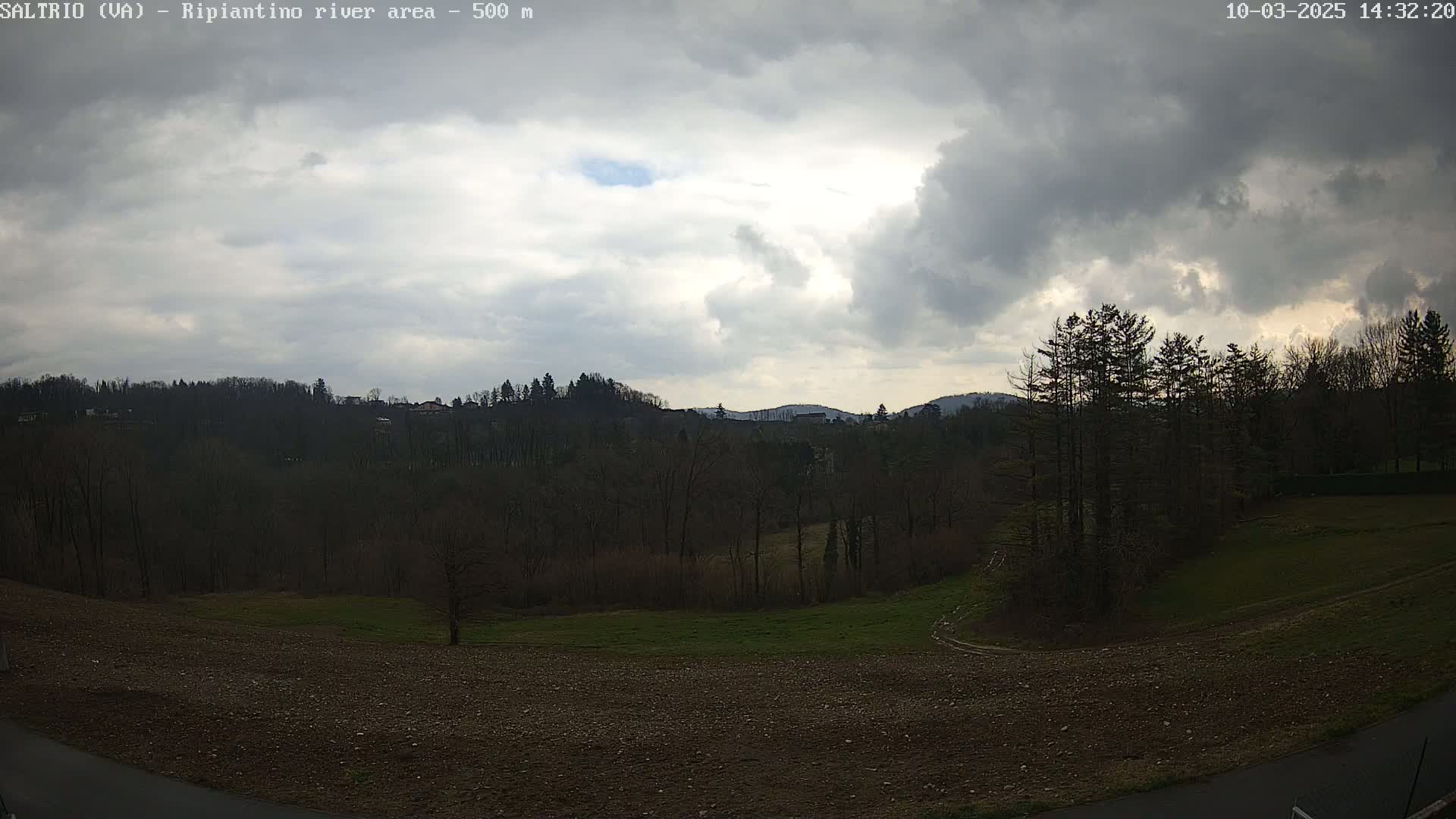 A mostly cloudy sky over a plowed field, bare trees, and distant hills.