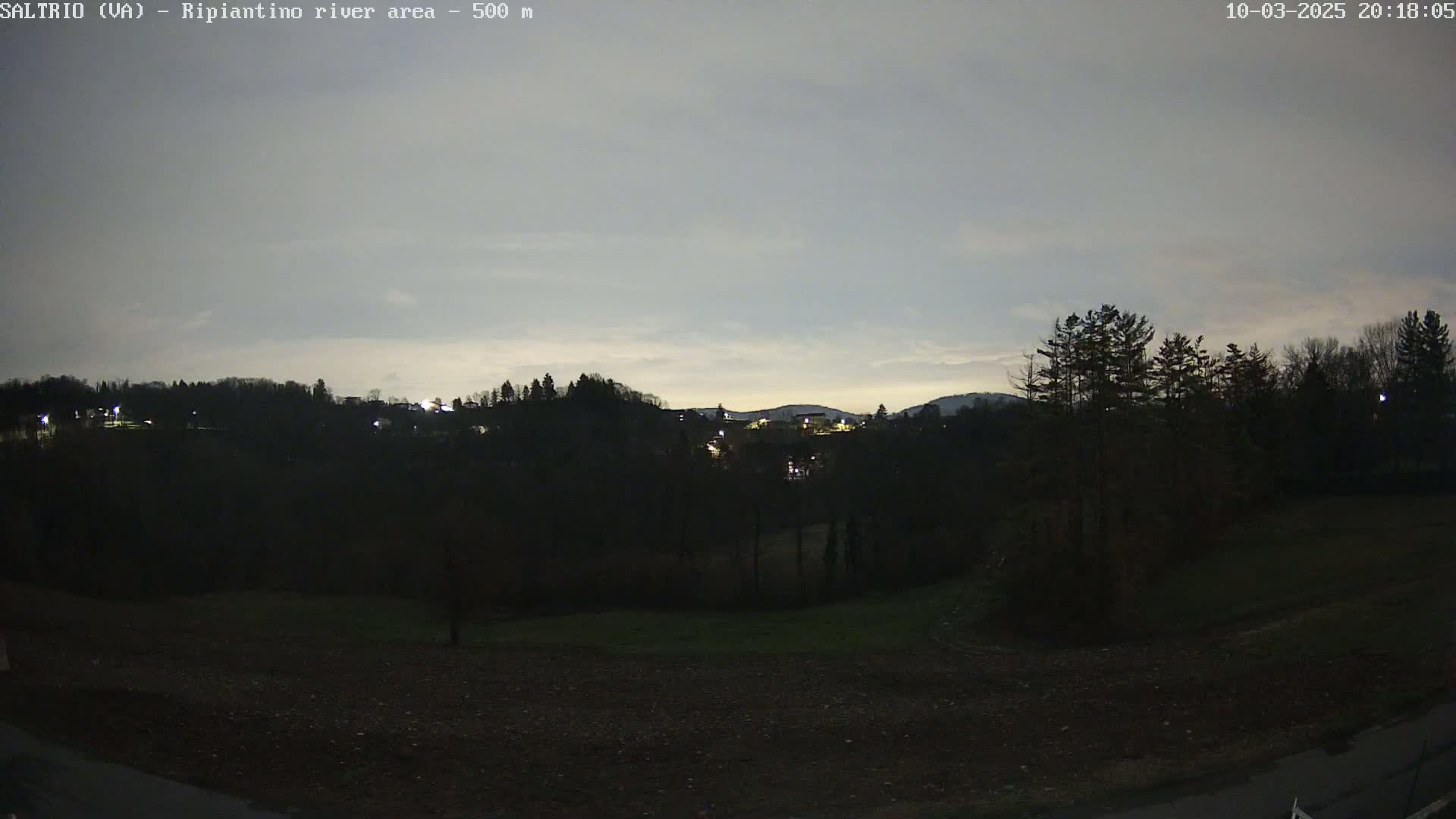 A nighttime view of a dark, hilly landscape with sparsely lit buildings in the distance under a twilight sky.