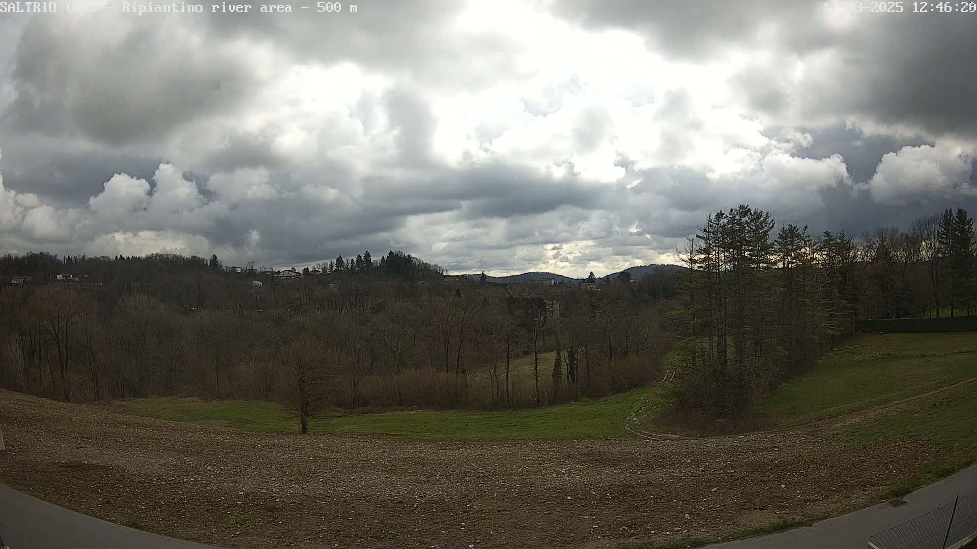 A mostly cloudy sky overlooks a landscape of bare winter trees, grassy fields, and tilled earth, with distant hills visible in the background.
