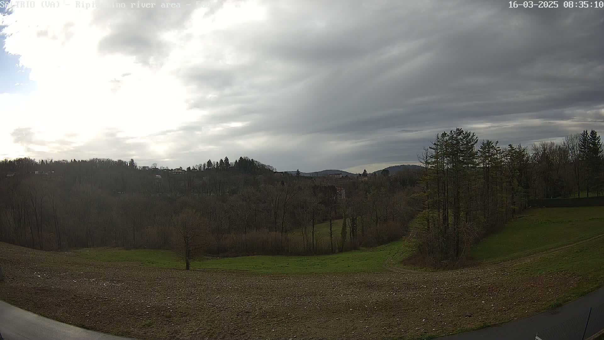 A mostly cloudy sky hangs over a hilly landscape of leafless trees, plowed fields, and a small patch of green grass.