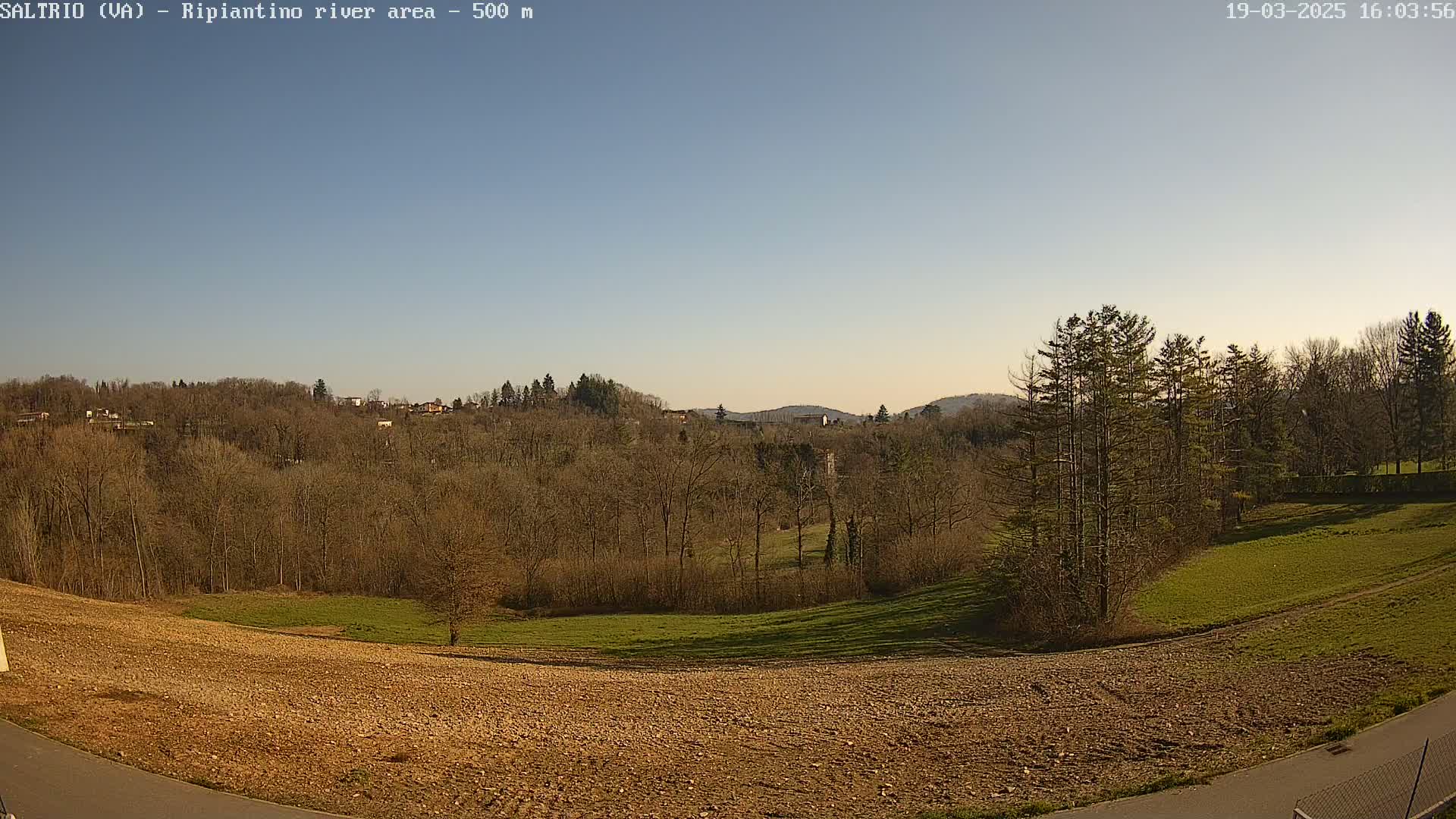 Under a clear sky, a plowed field slopes down toward a stand of leafless trees and a distant line of buildings nestled among more trees on a hillside.