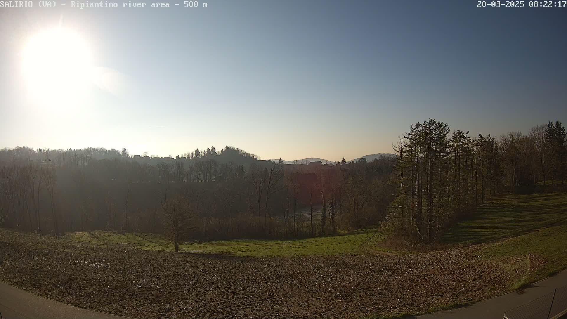 A sunny day reveals a view of a plowed field, grassy areas, and a line of trees, beyond which lies a river and hills covered in more trees.
