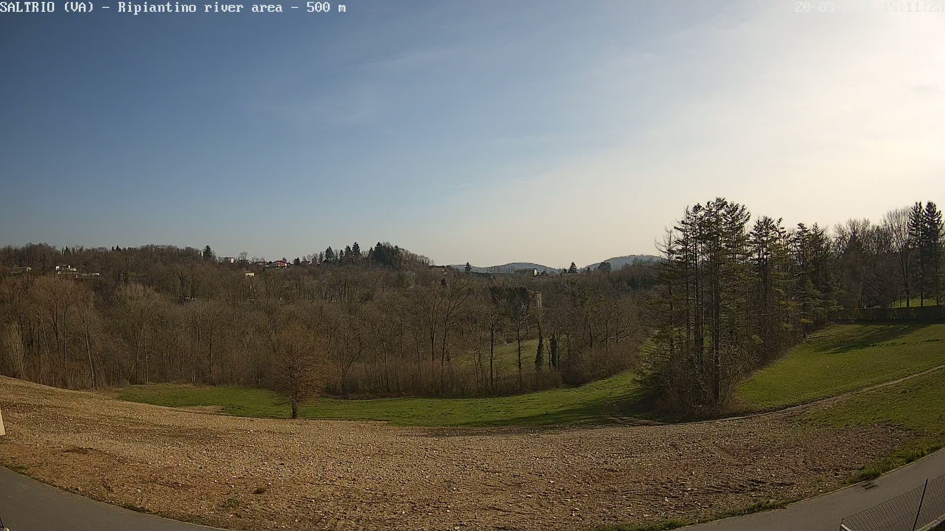A mostly barren field slopes down to a line of trees and distant hills under a clear blue sky.