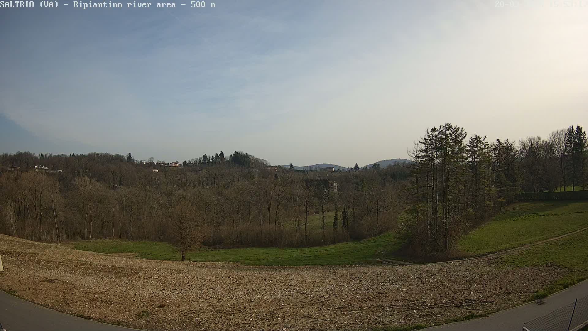 A mostly sunny day reveals a view of bare trees and rolling hills, with a grassy field and a rocky, plowed area in the foreground.