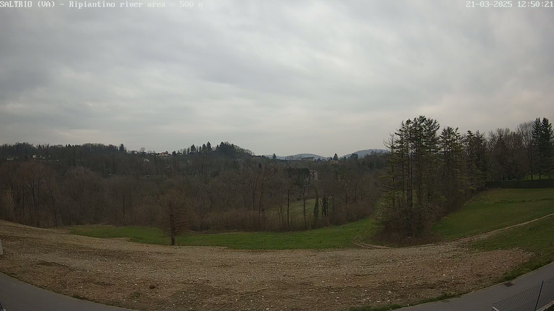 A view of a hilly landscape under a cloudy sky shows bare trees and a field of brown earth in the foreground.