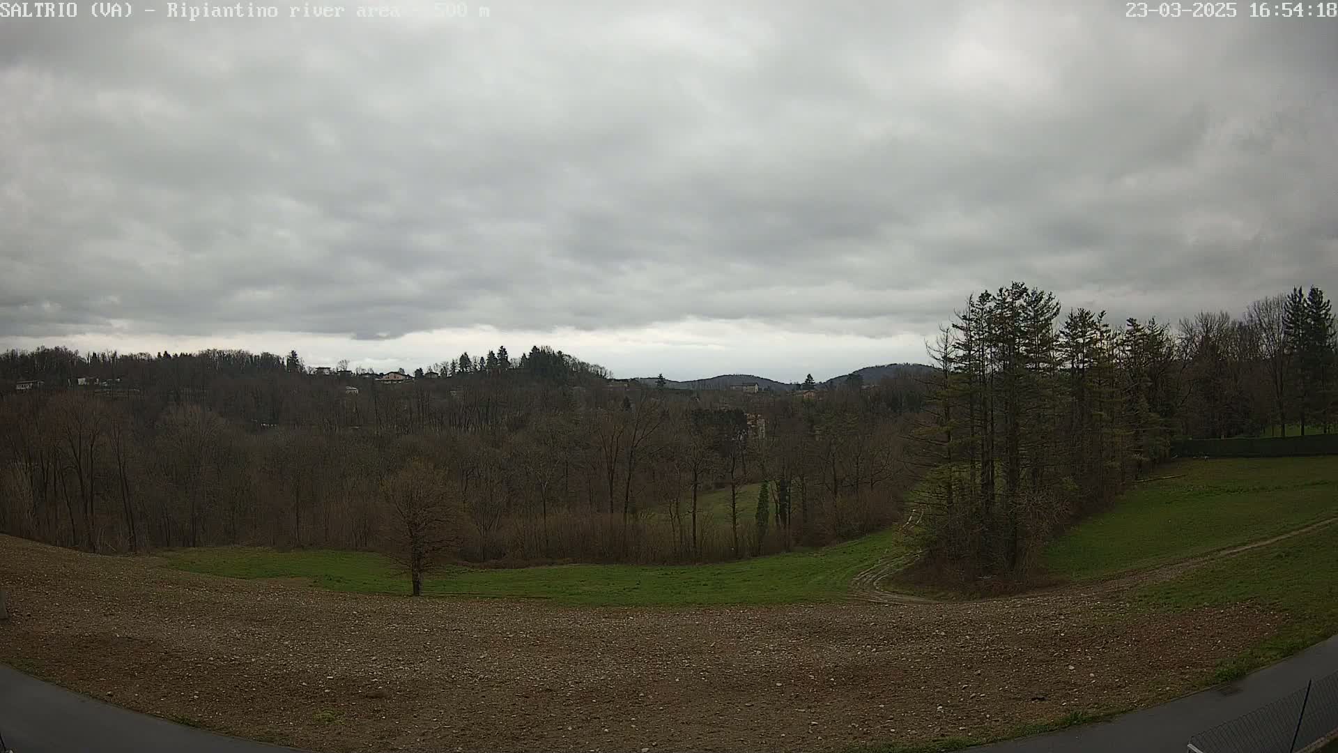 A mostly cloudy day reveals a view of a hilly landscape with bare trees and fields, some of which are tilled.