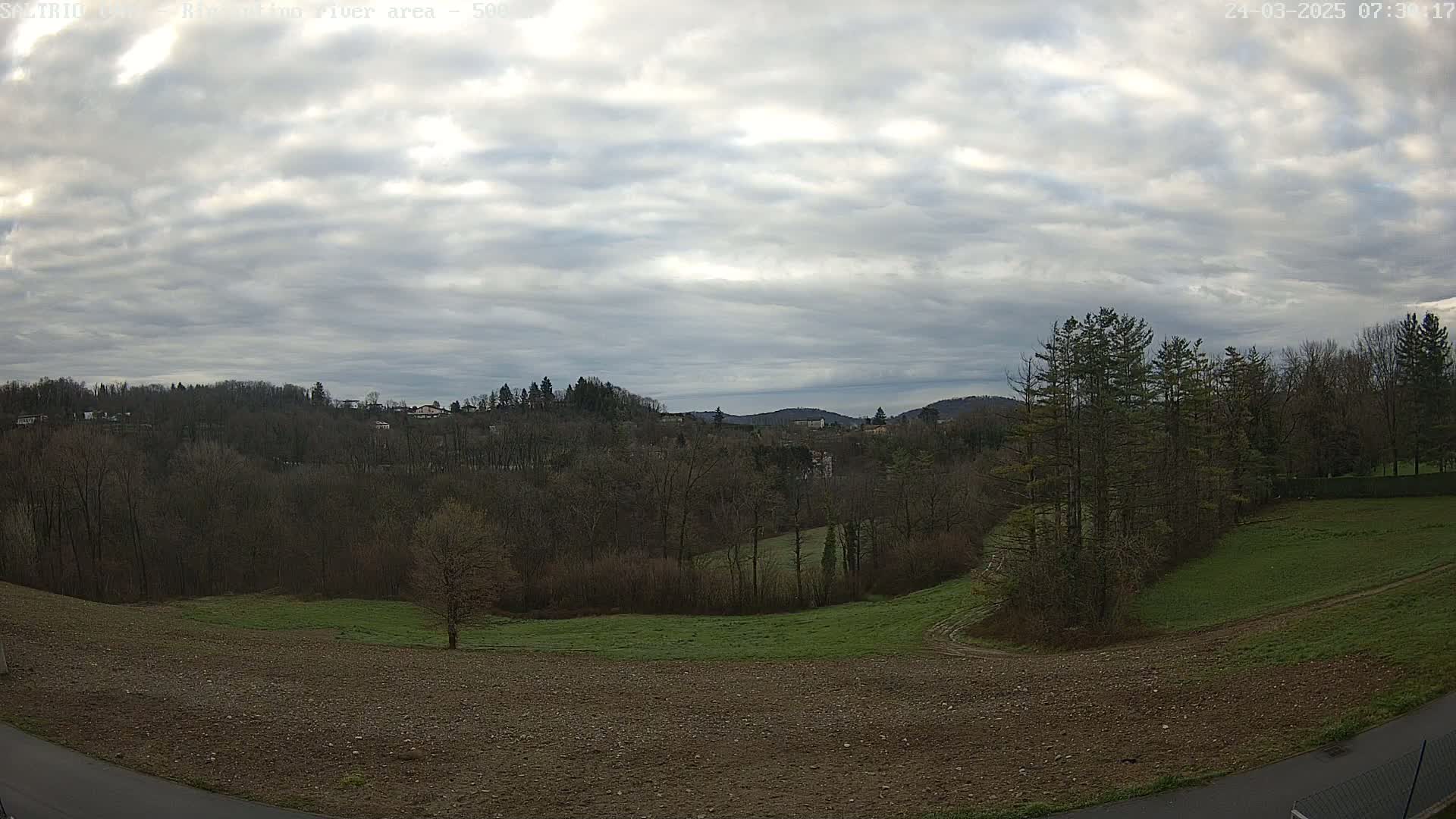 A view of a mostly cloudy sky over a landscape of bare winter trees, green and brown fields, and a few distant houses.