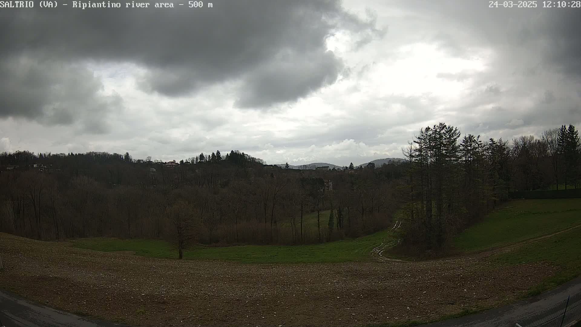A mostly cloudy sky overlooks a hilly landscape of bare trees, grassy fields, and a sparsely populated area.