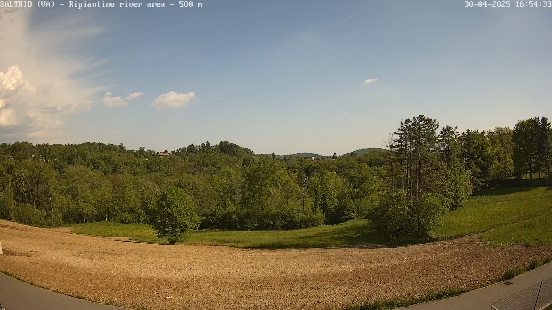 A mostly sunny day reveals a large, plowed field in the foreground, with a lush green forest and distant hills in the background.