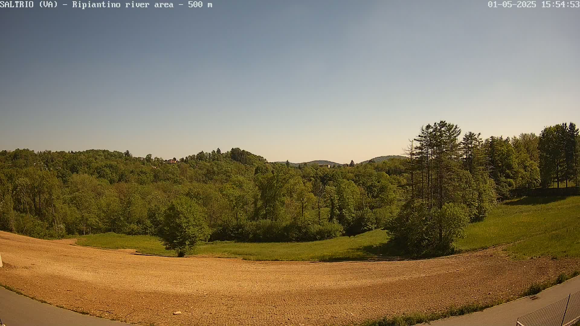A mostly clear sky overlooks a brown, plowed field next to a lush green forest and distant hills.