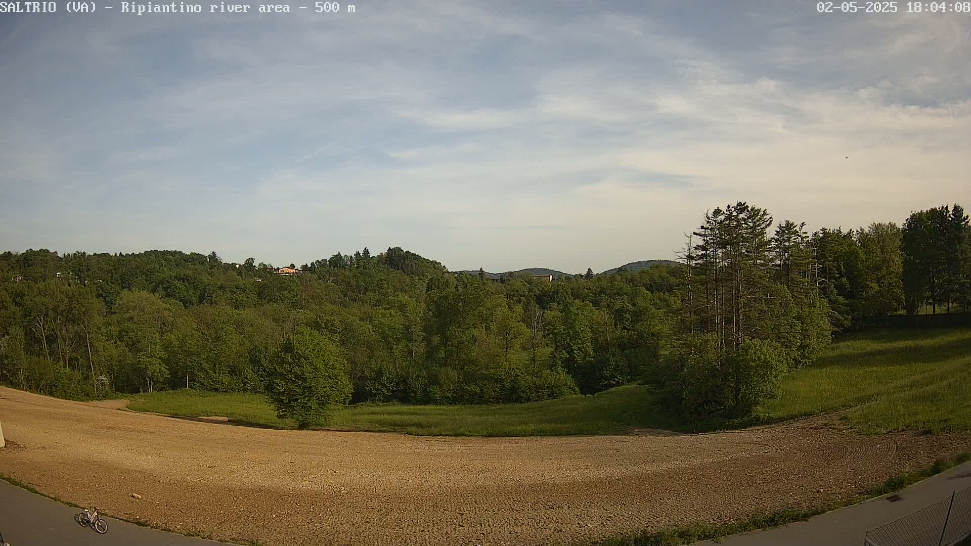 A mostly clear sky overlooks a mostly brown, barren field next to a lush green forest and a paved road with a bicycle on it.