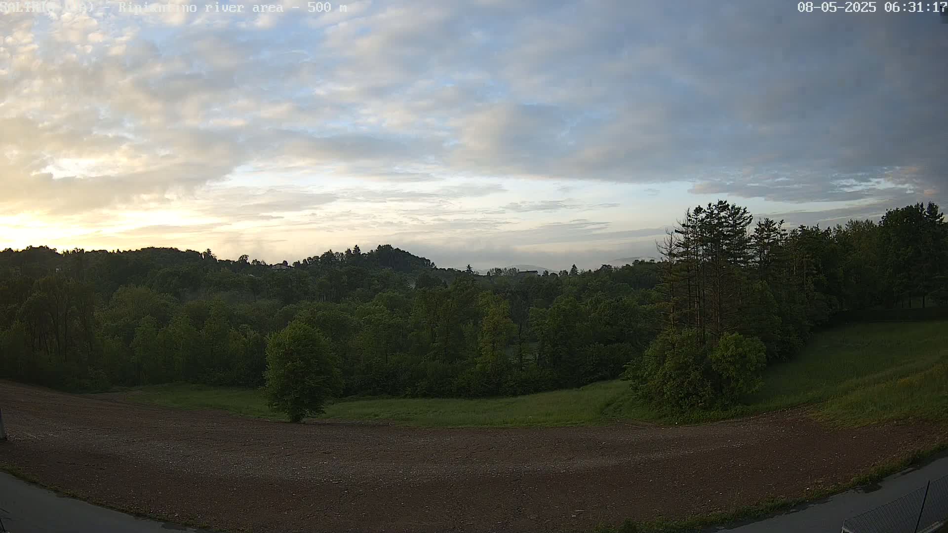 A mostly cloudy sunrise illuminates a verdant landscape of trees and a clearing with a brown, dirt path.
