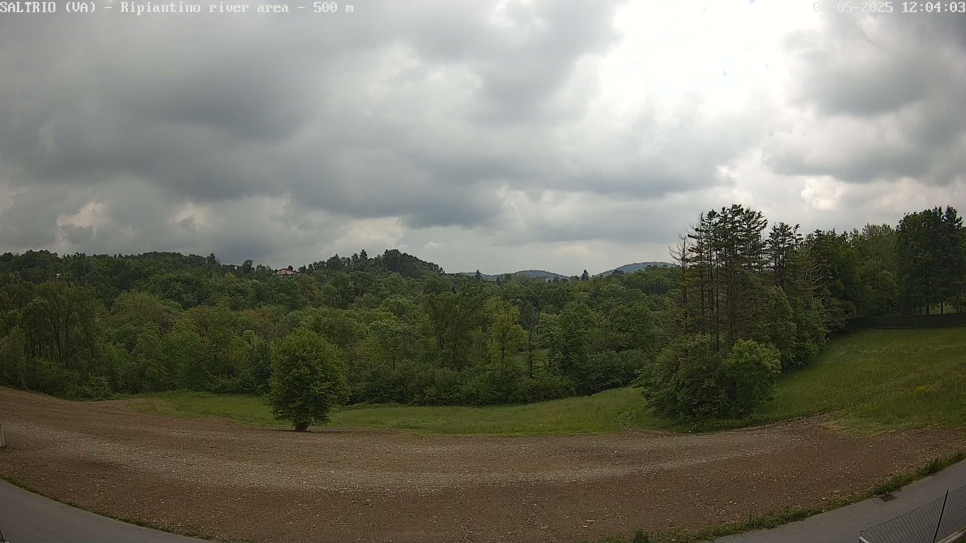 A mostly cloudy sky overlooks a largely wooded landscape with a cleared, gravelly area in the foreground.