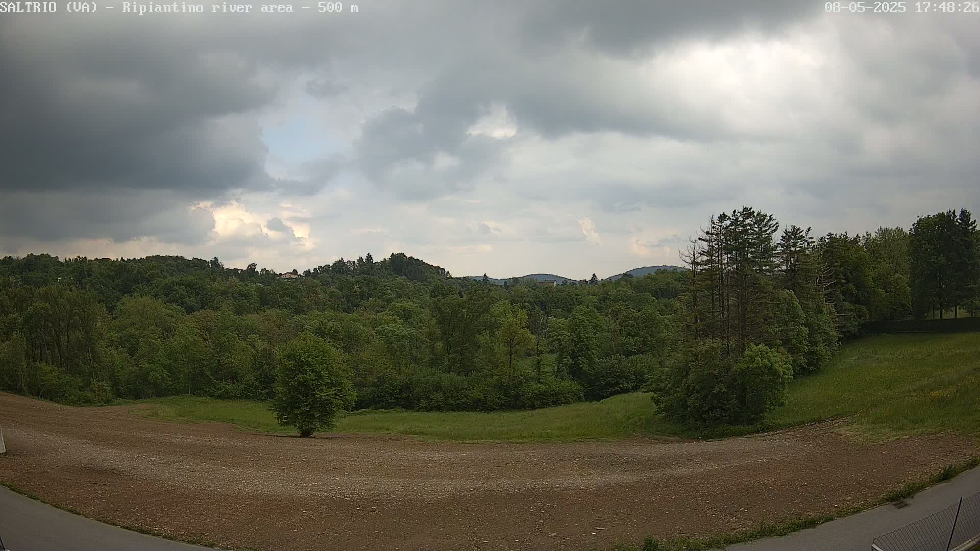 A mostly cloudy sky overlooks a verdant landscape of trees and fields, with a patch of bare, brown earth in the foreground.