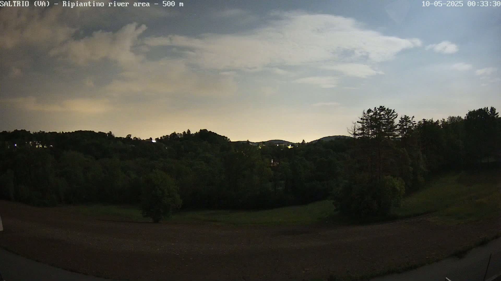 A dark, mostly cloudy night scene shows a hilly landscape covered in dark green trees and a foreground of brown, bare earth.