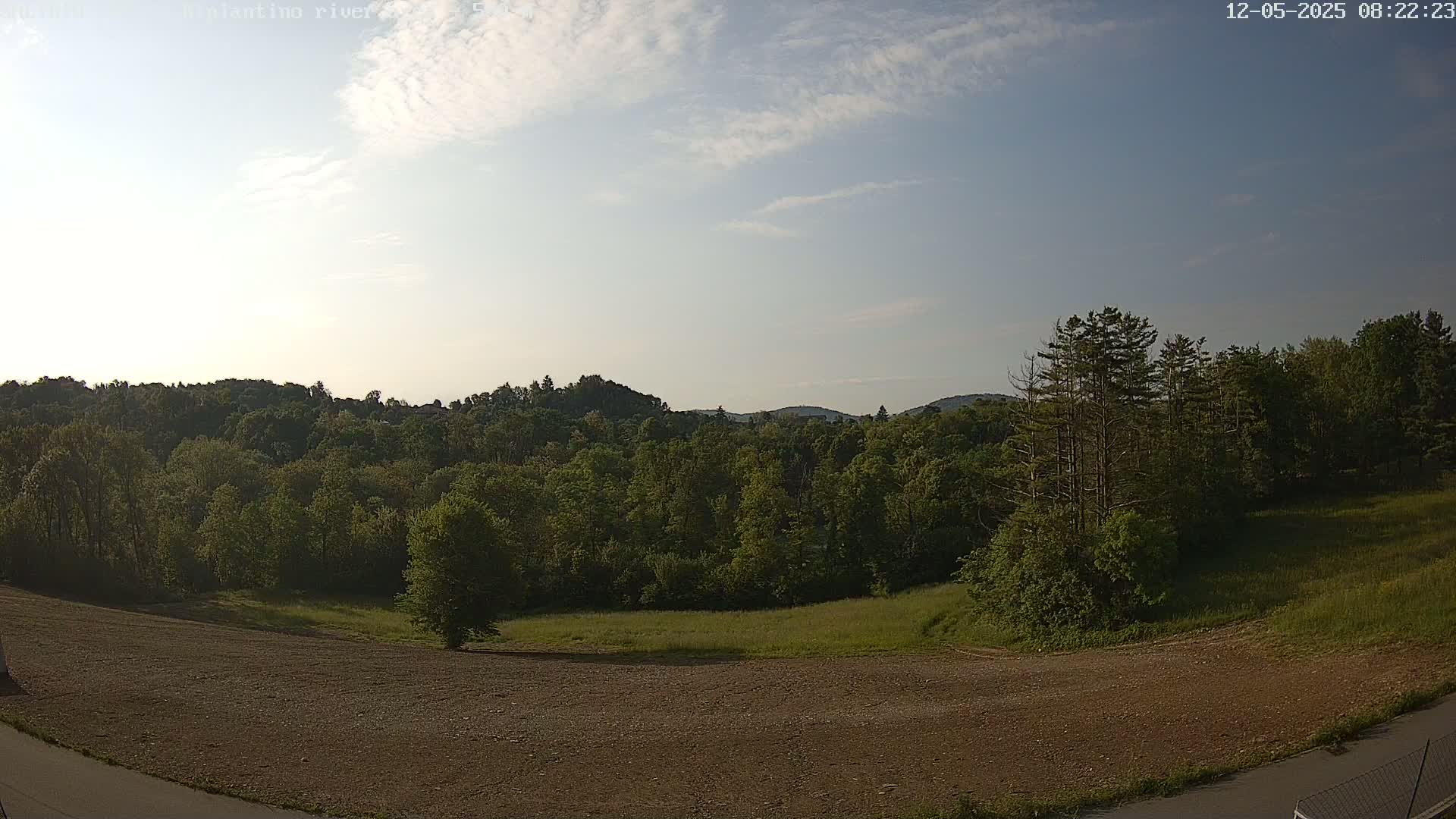 A mostly sunny day reveals a view of a verdant forest and hills, with a bare, brown patch of land in the foreground.