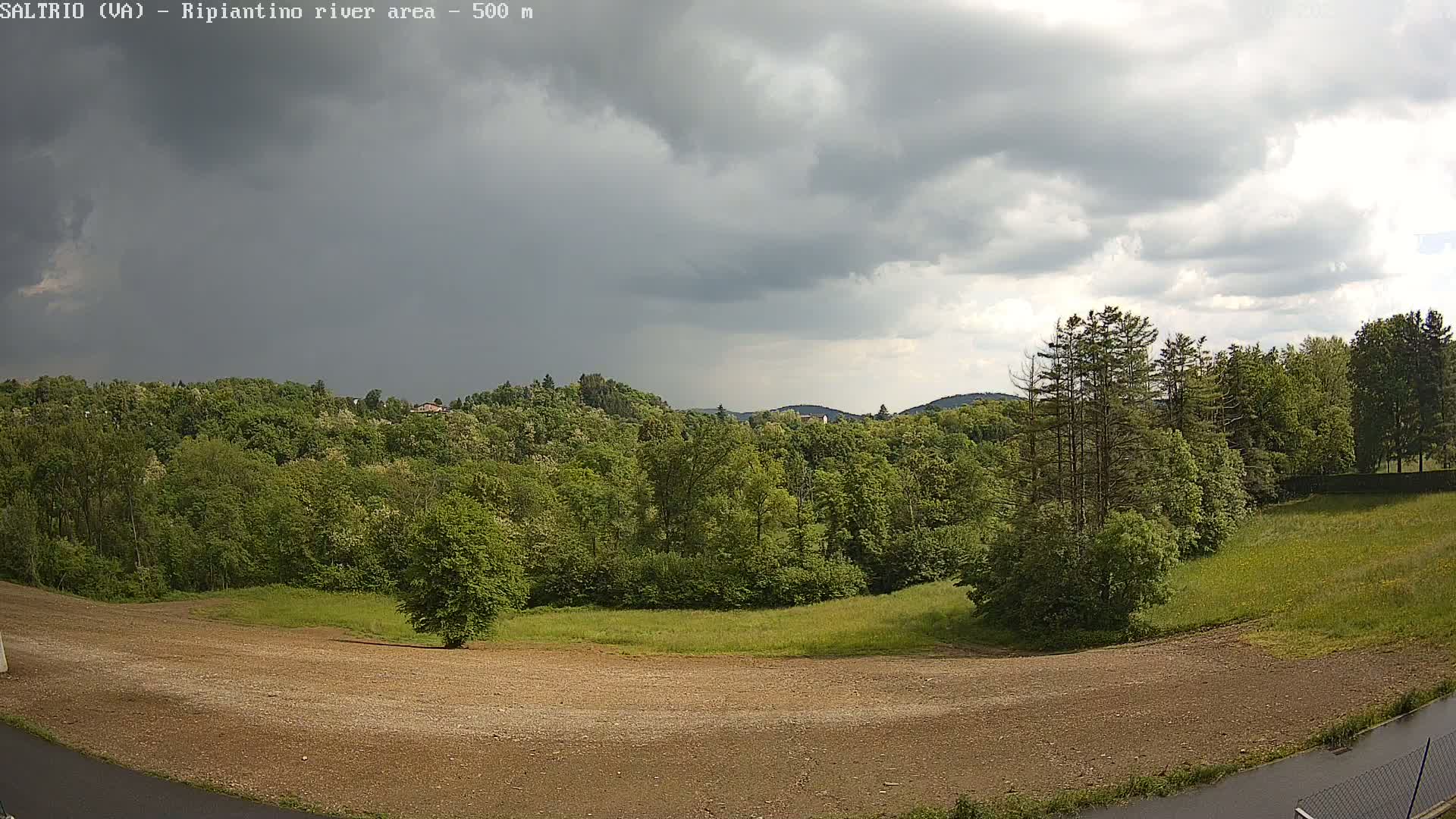 A mostly cloudy sky hangs over a landscape of lush green trees, a grassy field, and a large, bare patch of dirt.