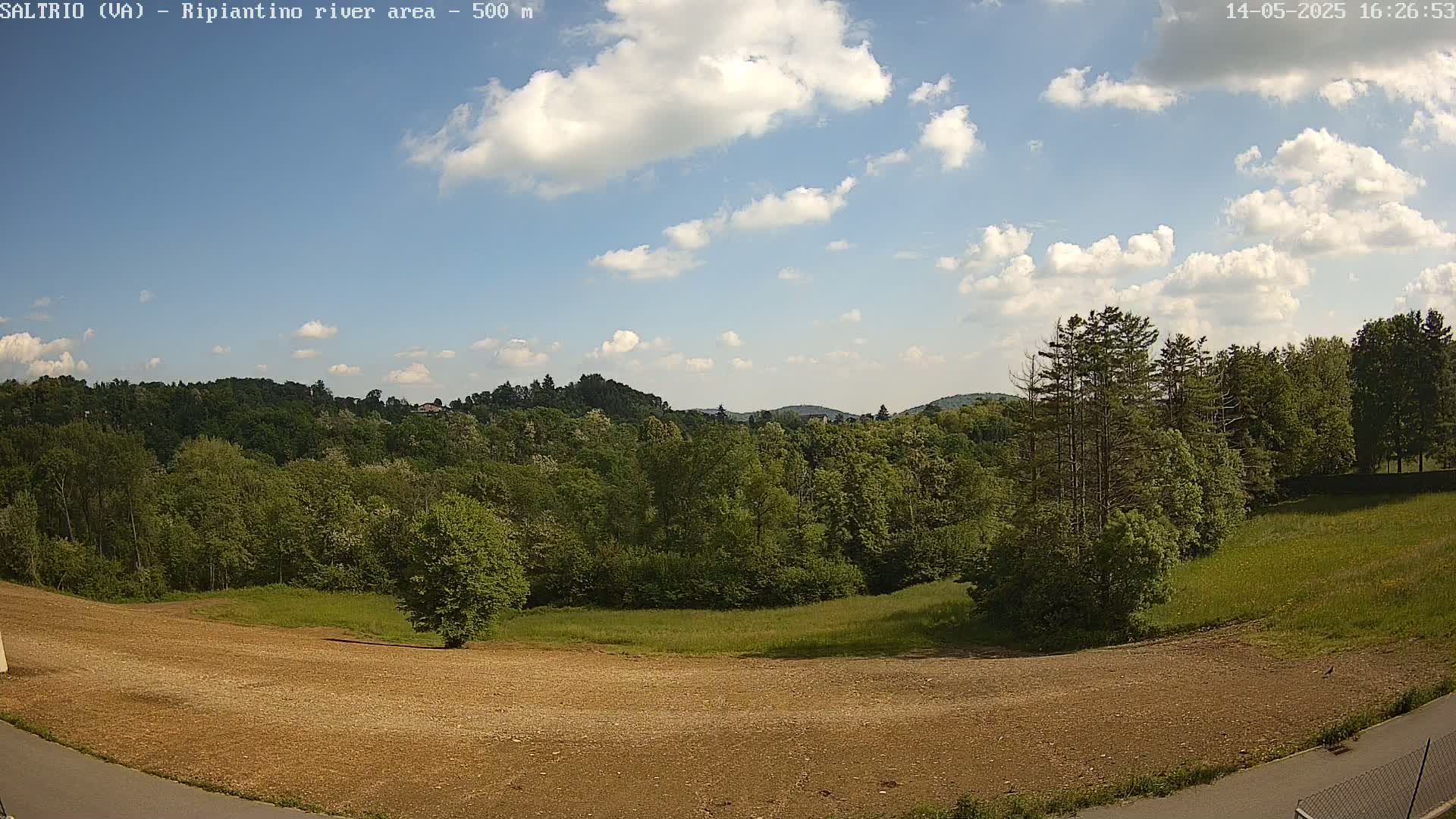 A mostly sunny day with scattered cumulus clouds over a landscape of lush green trees and a brown, cleared field.