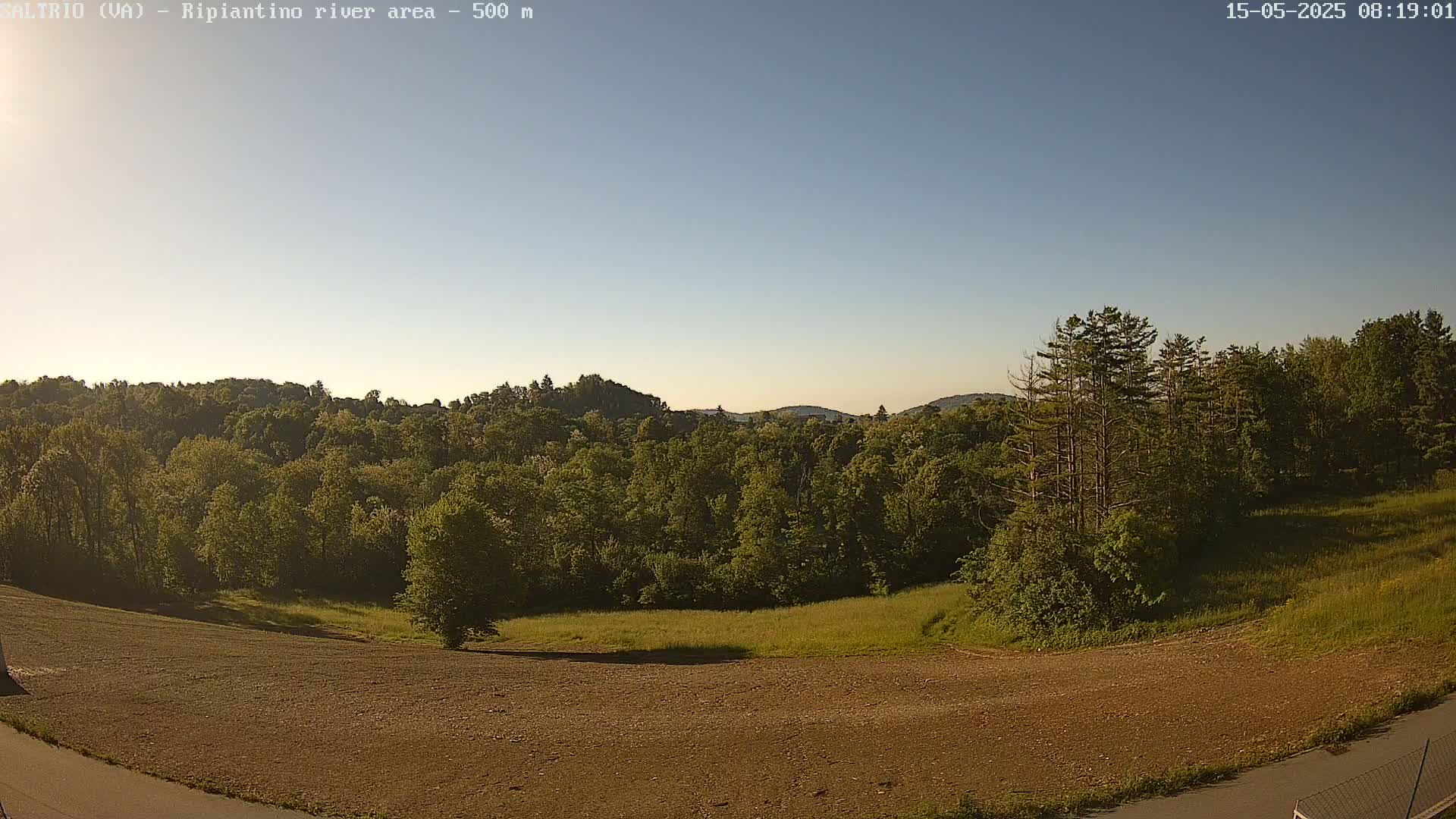 A mostly clear sky overlooks a plowed field, green grass, and a lush forest of deciduous trees on a hillside.