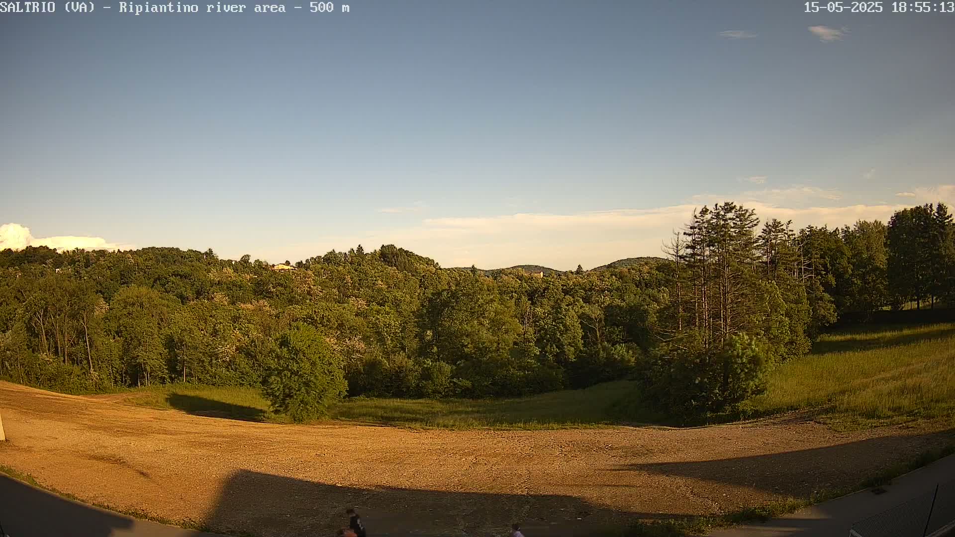 A clear, late afternoon view shows a mostly sunny sky over a large, flat, tan expanse of land, bordered by lush green trees and hills in the background.