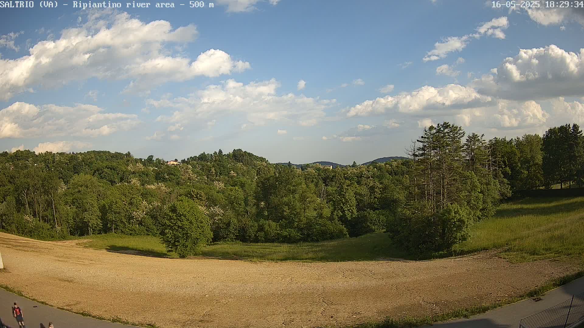 A partly sunny day reveals a view of a lush green forest and a large expanse of bare, light brown earth, with a few people visible in the bottom left corner.