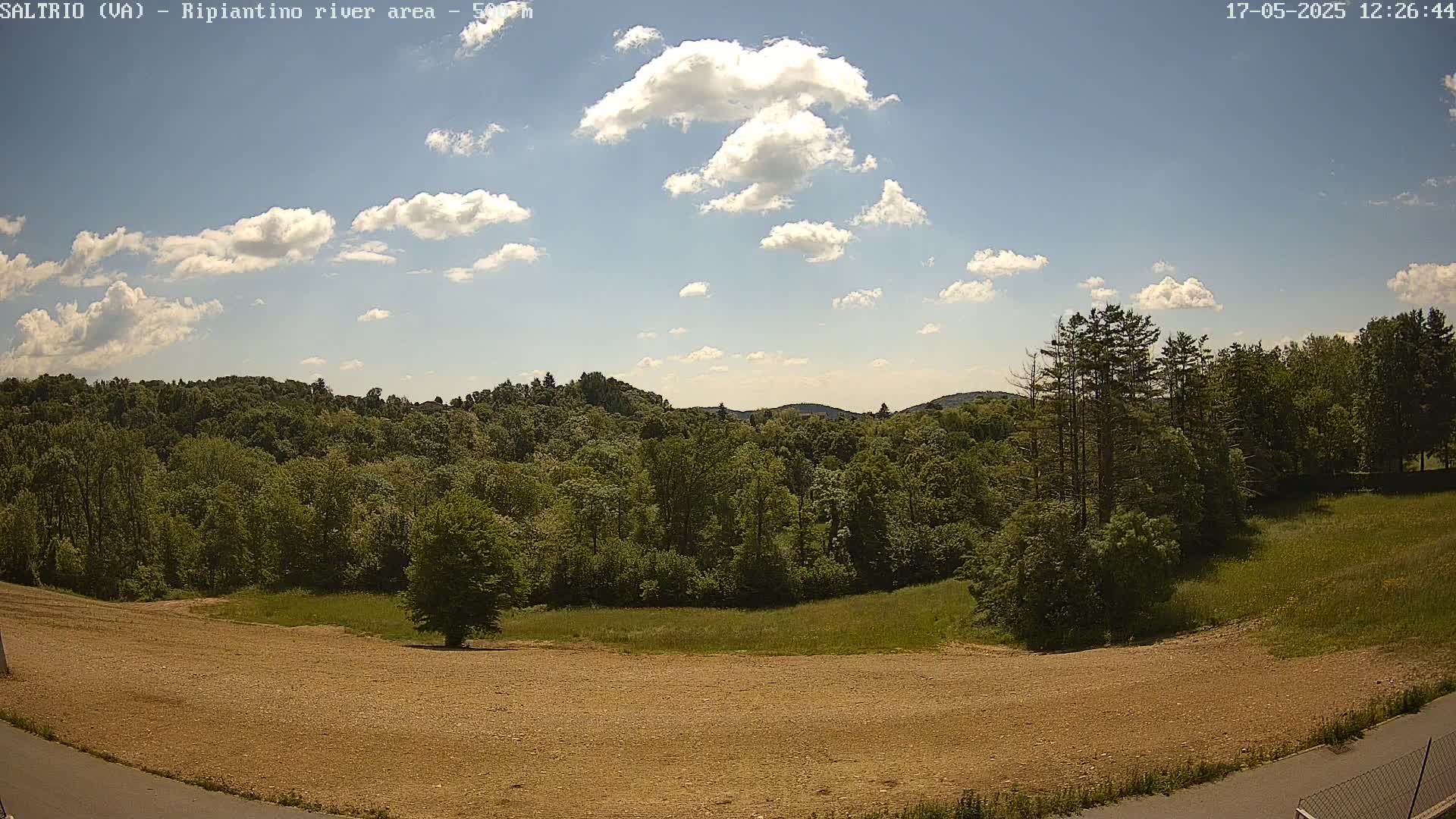 A mostly sunny sky with scattered cumulus clouds overlooks a verdant landscape of trees and a cleared, tan-colored field.
