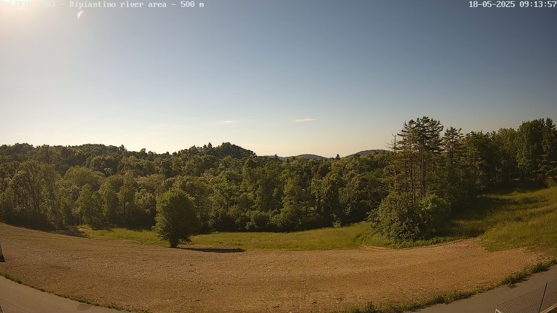 A mostly clear sky overlooks a plowed field, grassy areas, and a dense forest of green trees on a hillside.