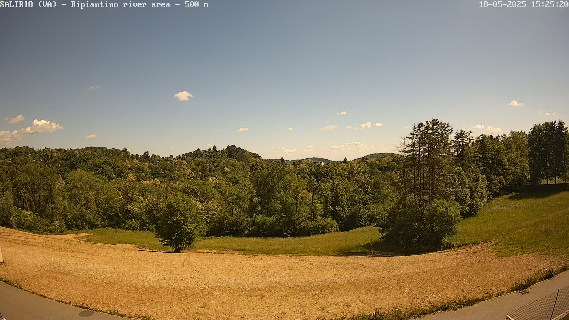 A mostly sunny day reveals a view of a plowed field, grassy areas, and lush green trees on a hillside, under a mostly clear sky.