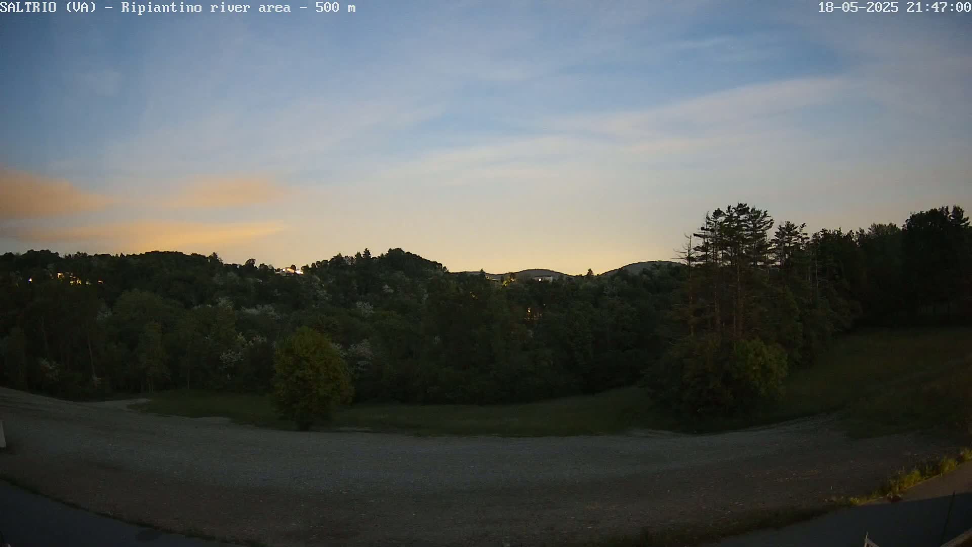 A dusky twilight scene shows a hill covered in dark trees, a flat, gravel area in the foreground, and a pale, orange-tinged sky.