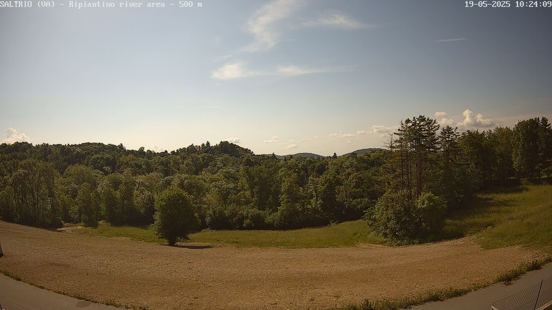 A mostly sunny day reveals a view of a large, grassy field sloping down to a paved road, bordered by a lush green forest that extends into rolling hills under a mostly clear sky.